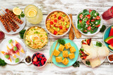 Summer food table scene over a white wood background. Collection of refreshing salads, fruit, wraps and BBQ grilled skewers. Top view.