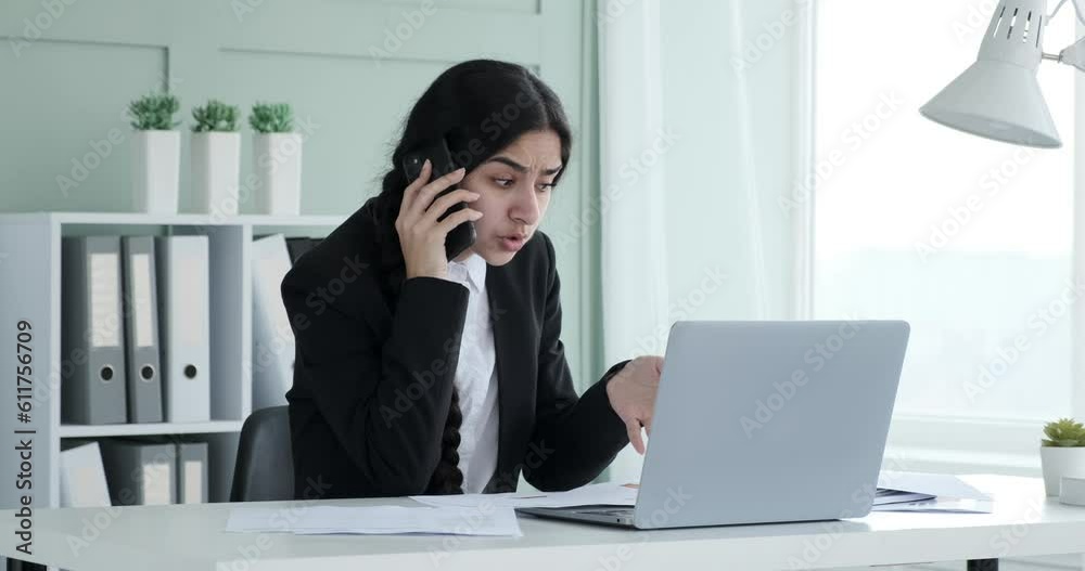 An Indian businesswoman sits at office desk, engrossed in a phone conversation. As the call ends, she sets the phone on the desk and expresses exasperation by clutching her head with a sigh of relief. An Indian businesswoman sits at office desk, engrossed in a phone conversation. As the call ends, she sets the phone on the desk and expresses exasperation by clutching her head with a sigh of relief.