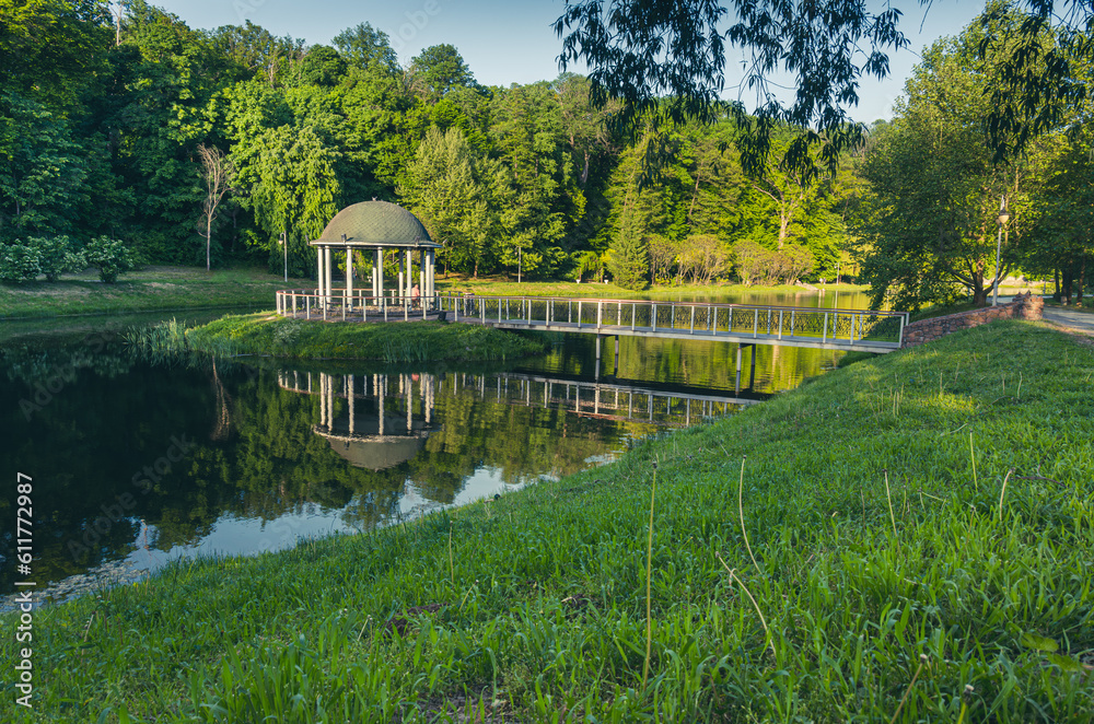 An alcove on the water, a bridge leading to it, green grass and a lake in the foreground