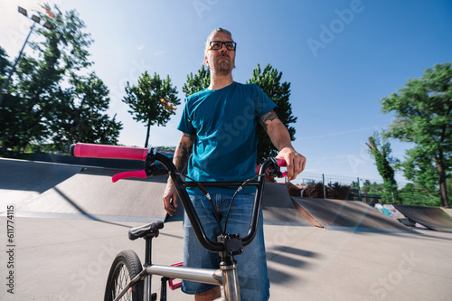Portrait of an oldish tattooed guy posing with his bmx in a skate park.