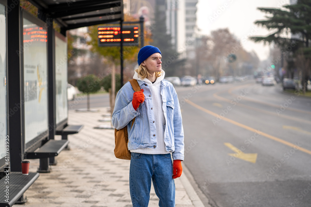 Young guy tourist with backpack waiting at bus stop early in morning. Male passenger use public transport in city. Move without car in big city. Teen waiting bus very long time and looks into distance Young guy tourist with backpack waiting at bus stop early in morning. Male passenger use public transport in city. Move without car in big city. Teen waiting bus very long time and looks into distance