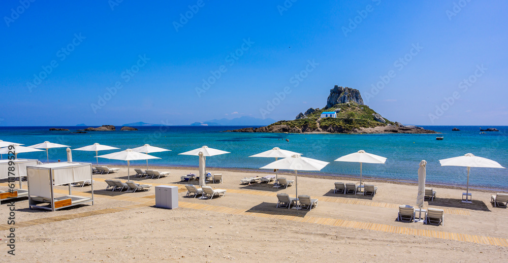 Deck chair and umbrella on beautiful Agios Stefanos Beach in front of paradise Island Kastri- historical ruins and paradise scenery at coast of island Kos, Greece