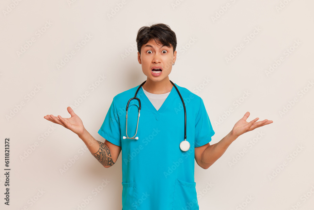 Young male resident doctor in turquoise medical uniform with stethoscope on his neck stands with his mouth open and spreading arms sideways, health care concept, copy space Young male resident doctor in turquoise medical uniform with stethoscope on his neck stands with his mouth open and spreading arms sideways, health care concept, copy space