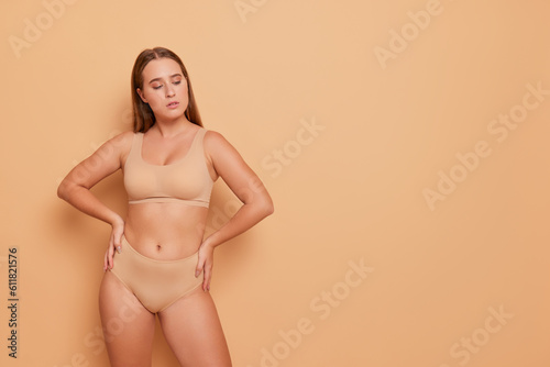 Pretty girl in beige underwear standing against background in studio with arms akimbo, looking down, comfortable underwear concept, copy space