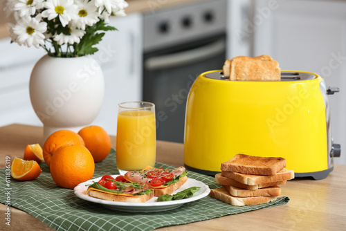 Modern toaster with crispy bread slices, glass of juice, orange and delicious sandwiches on table in kitchen
