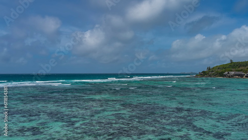 The waves are foaming over the reefs in the boundless turquoise ocean. On the shore of the island, the villas of the hotel are visible in the distance. Blue sky, clouds. Seychelles. Moyenne Island.