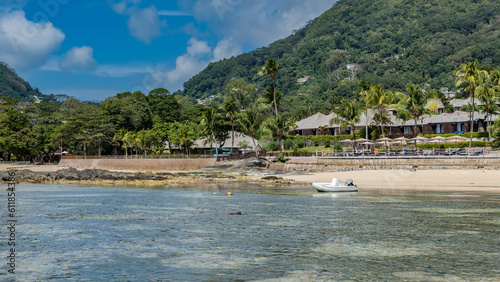 Low tide on a tropical beach. The rocks of the seabed were exposed. The white boat is moored in shallow water. On the shore the sun umbrellas, the villas of the hotel are visible. Seychelles. Mahe.