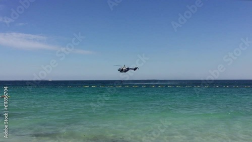 Helicopter lowers rescuer just above aquamarine water while training. It moves slowly forward beneath near clear blue sky. Swimmer in foreground