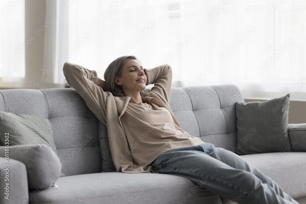 Peaceful happy grey haired homeowner woman enjoying leisure, weekend at home, sitting on comfortable couch, leaning on back, taking deep breath with closed eyes, smiling
