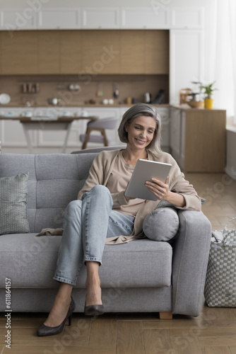 Cheerful positive pretty grey haired woman holding digital gadget, enjoying leisure, communication on comfortable couch, using application on tablet computer. Vertical full length shot