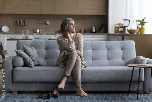 Tired serious middle aged grey haired woman sitting on home couch after long business day, taking shoes off, touching neck, looking away, thinking, feeling fatigue, burnout