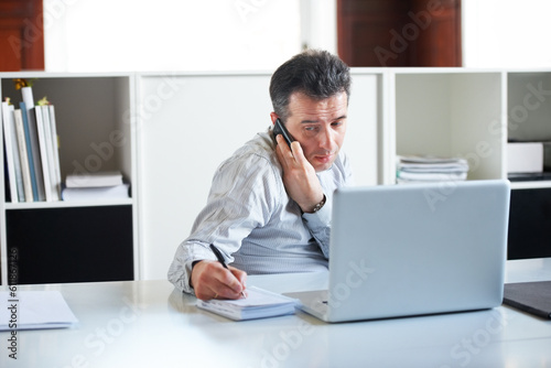 Consultant, businessman with smartphone on a call and with laptop writing notes in a notebook at his office. Customer service or online communication, networking and male person a cellphone at desk