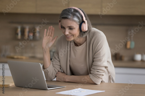Happy mature freelance business woman in big wireless headphones talking on video conference call, waving hand hello, speaking, smiling, enjoying Internet communication with colleagues, teacher