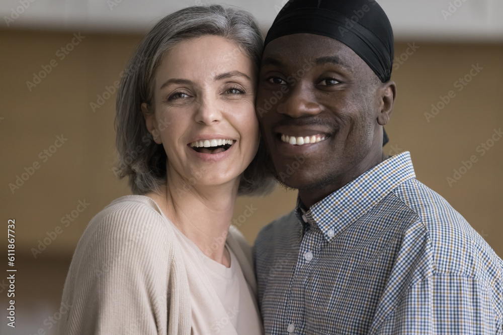 Happy diverse African and Caucasian couple posing indoors, standing close with cheeks touch, looking away with toothy smiles, expressing love, promoting warm healthy relationship. Casual portrait