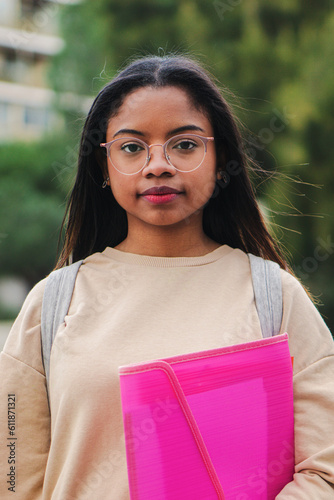 Vertical portrait of smart young latin school girl holding the notebook with pensive attitude. Serious hispanic teenage female standing outdoors at university capus looking at camera with his googles photo