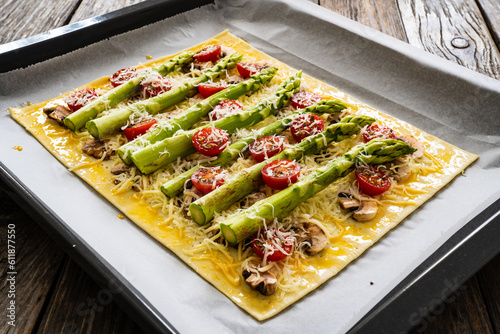 Uncooked puff pastry, green asparagus with mozzarella cheese, mushrooms and cherry tomatoes on baking paper on cooking pan
