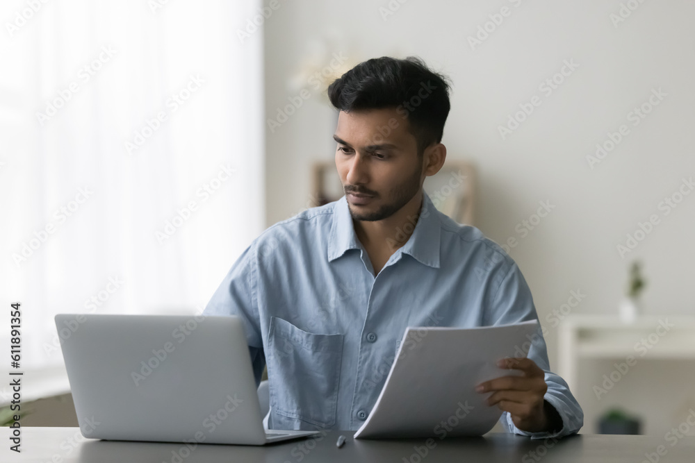 Serious young Indian entrepreneur man reviewing paper documents at work table, working at laptop, looking at screen, checking contract, agreement, consulting online legal service Serious young Indian entrepreneur man reviewing paper documents at work table, working at laptop, looking at screen, checking contract, agreement, consulting online legal service