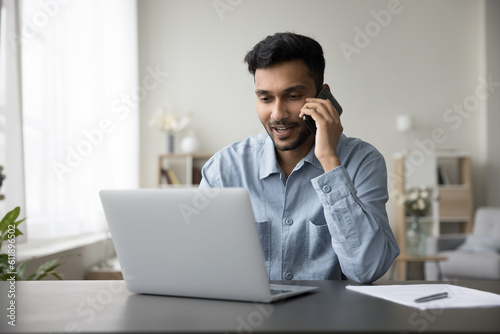 Positive Indian freelance professional man talking on cellphone at laptop, working on business project at home, enjoying online communication, using Internet technology for job