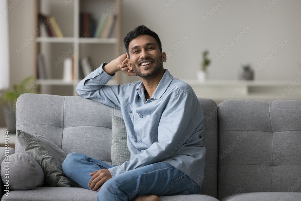 Cheerful handsome Indian guy relaxing on comfortable sofa at home, leaning hand on soft on back, enjoying comfort, leisure, looking at camera, smiling, laughing Cheerful handsome Indian guy relaxing on comfortable sofa at home, leaning hand on soft on back, enjoying comfort, leisure, looking at camera, smiling, laughing