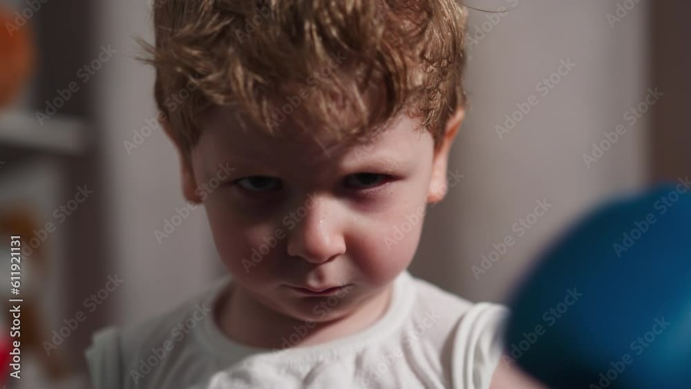 Active little boy waves with boxing glove on hand and frowns looking ahead. Sportive redhead child in t-shirt wants to scare opponent on ring closeup slow motion Active little boy waves with boxing glove on hand and frowns looking ahead. Sportive redhead child in t-shirt wants to scare opponent on ring closeup slow motion