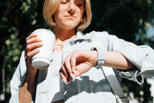 Close-up of young stylish caucasian woman in glasses and with cup of coffee looking at an electronic wristwatch outdoors, lifestyle. Selective focus on female hand with device