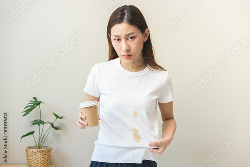 Cloth stain, disappointment asian young woman clumsy with hot coffee, tea stains on shirt, hand show making spill drop on white t-shirt, spot dirty or smudge on clothes at home, isolated on background photo