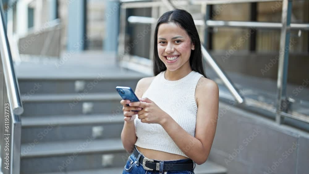 Young beautiful hispanic woman using smartphone smiling at street