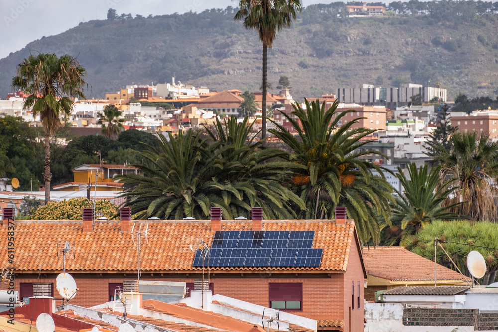 Solar panels on the roof of a single family house of red bricks and red tiles, with palm trees and landscape of trees in the background. Tenerife, Canary Islands, Spain