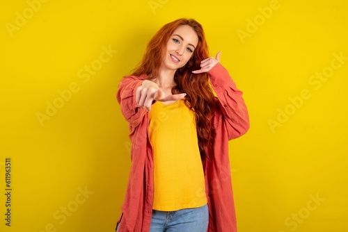 Young redhead woman wearing fashion clothing over yellow wall smiling cheerfully and pointing to camera while making a call you later gesture, talking on phone
