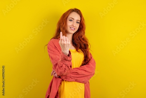 Young redhead woman wearing fashion clothing over yellow wall Beckoning come here gesture with hand inviting welcoming happy and smiling