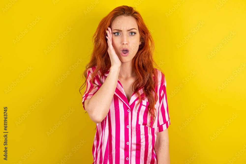 Shocked Young red haired woman wearing pink pyjama over yellow studio background looks with great surprisment being very stunned, astonished with unexpected news, Facial expressions concept. Shocked Young red haired woman wearing pink pyjama over yellow studio background looks with great surprisment being very stunned, astonished with unexpected news, Facial expressions concept.