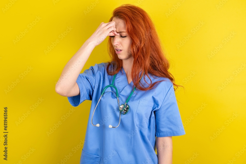 Sad redhead doctors woman wearing blue uniform over yellow wall suffering from headache holding hand on her face Sad redhead doctors woman wearing blue uniform over yellow wall suffering from headache holding hand on her face