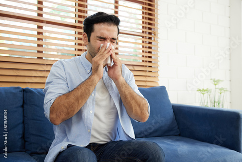 young man having a cold, using tissue paper and blowing his nose on sofa