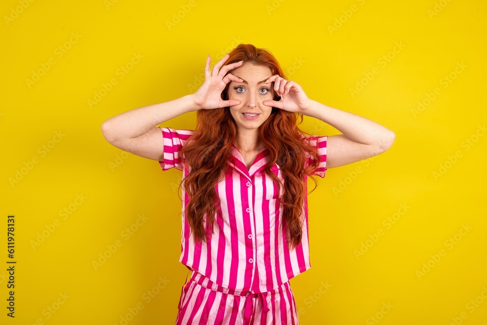 Young red haired woman wearing pink pyjama over yellow studio background keeping eyes opened to find a success opportunity.