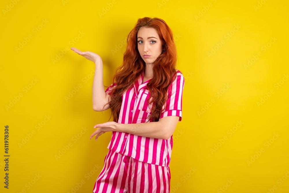 Young red haired woman wearing pink pyjama over yellow studio background pointing aside with ...