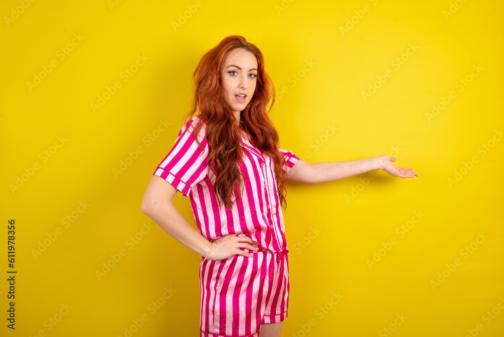 Young red haired woman wearing pink pyjama over yellow studio background feeling happy and cheerful, smiling and welcoming you, inviting you in with a friendly gesture Young red haired woman wearing pink pyjama over yellow studio background feeling happy and cheerful, smiling and welcoming you, inviting you in with a friendly gesture