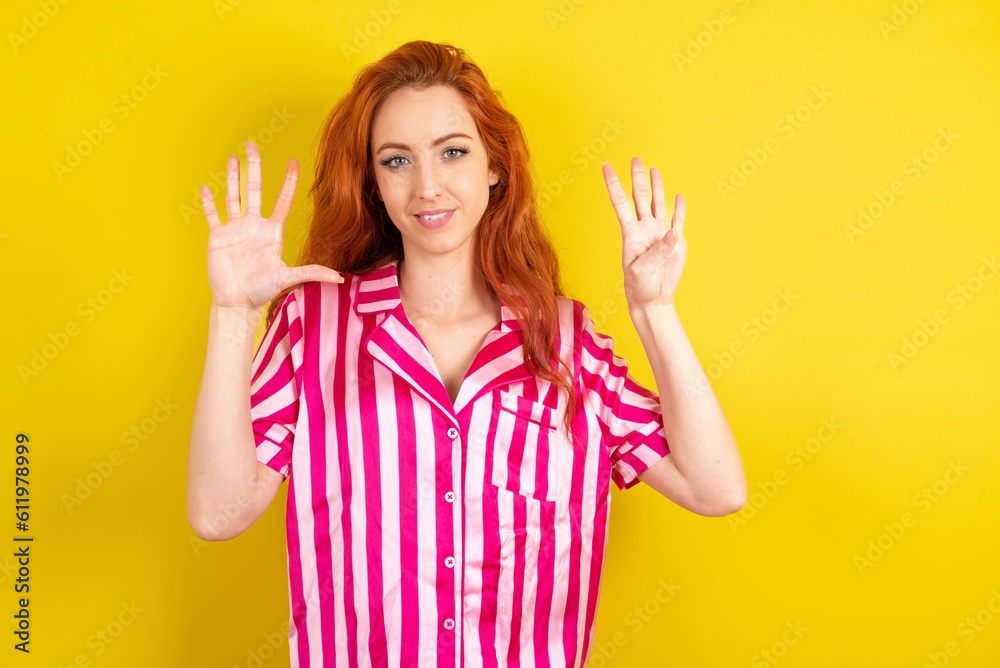 Young red haired woman wearing pink pyjama over yellow studio background showing and pointing up with fingers number nine while smiling confident and happy. Young red haired woman wearing pink pyjama over yellow studio background showing and pointing up with fingers number nine while smiling confident and happy.