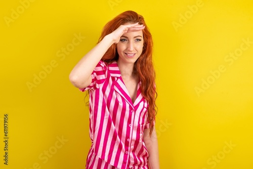 Young red haired woman wearing pink pyjama over yellow studio background very happy and smiling looking far away with hand over head. Searching concept.