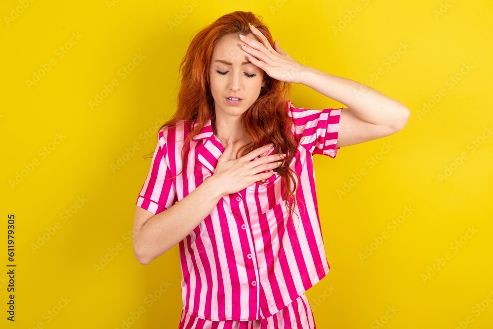 Young red haired woman wearing pink pyjama over yellow studio background Touching forehead for illness and fever, flu and cold, virus sick.