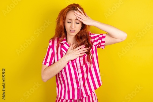 Young red haired woman wearing pink pyjama over yellow studio background Touching forehead for illness and fever, flu and cold, virus sick.