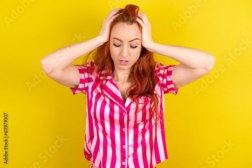 Young red haired woman wearing pink pyjama over yellow studio background suffering from strong headache desperate and stressed because of overwork. Depression and pain concept.