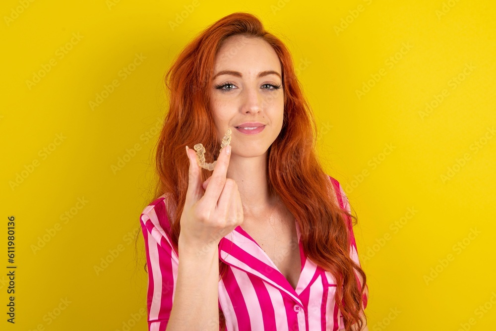 Young red haired woman wearing pink pyjama over yellow studio background holding an invisible aligner ready to use it. Dental healthcare and confidence concept. Young red haired woman wearing pink pyjama over yellow studio background holding an invisible aligner ready to use it. Dental healthcare and confidence concept.