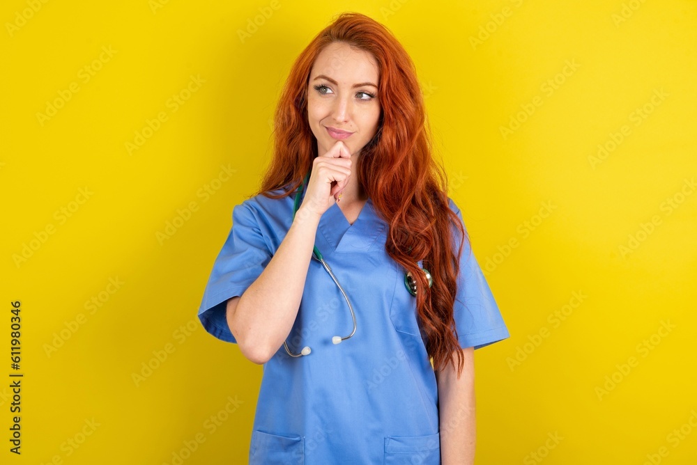 Thoughtful young red-haired doctor woman over yellow studio background holds chin and looks away pensively makes up great plan Thoughtful young red-haired doctor woman over yellow studio background holds chin and looks away pensively makes up great plan