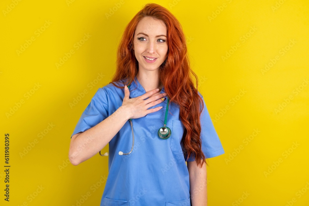 Joyful young red-haired doctor woman over yellow studio background expresses positive emotions recalls something funny keeps hand on chest and giggles happily. Joyful young red-haired doctor woman over yellow studio background expresses positive emotions recalls something funny keeps hand on chest and giggles happily.