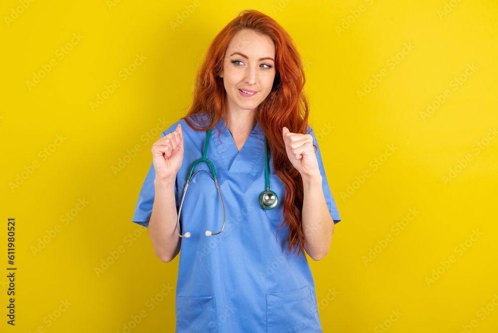 young red-haired doctor woman over yellow studio background clenches fists and awaits for something nice happened looks away bites lips and waits announcement of results young red-haired doctor woman over yellow studio background clenches fists and awaits for something nice happened looks away bites lips and waits announcement of results