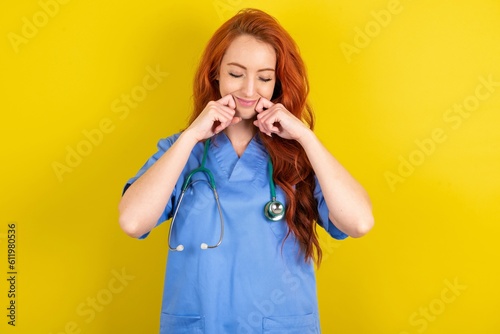 Pleased young red-haired doctor woman over yellow studio background with closed eyes keeps hands near cheeks and smiles tenderly imagines something very pleasant