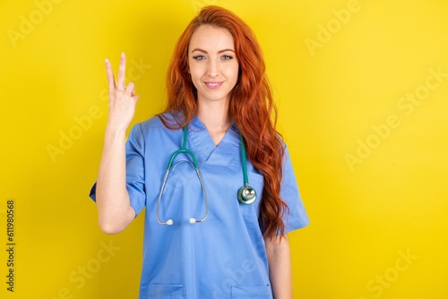 young red-haired doctor woman over yellow studio background  smiling and looking friendly, showing number two or second with hand forward, counting down