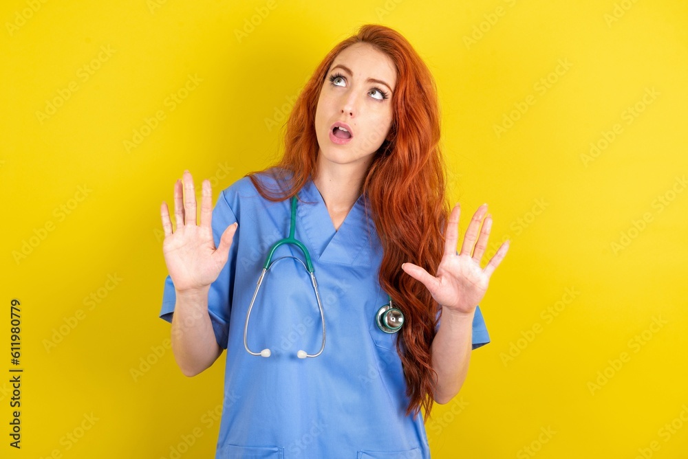 young red-haired doctor woman over yellow studio background keeps palms forward and looks with fright above on ceiling tries to defense herself from invisible danger opens mouth. young red-haired doctor woman over yellow studio background keeps palms forward and looks with fright above on ceiling tries to defense herself from invisible danger opens mouth.