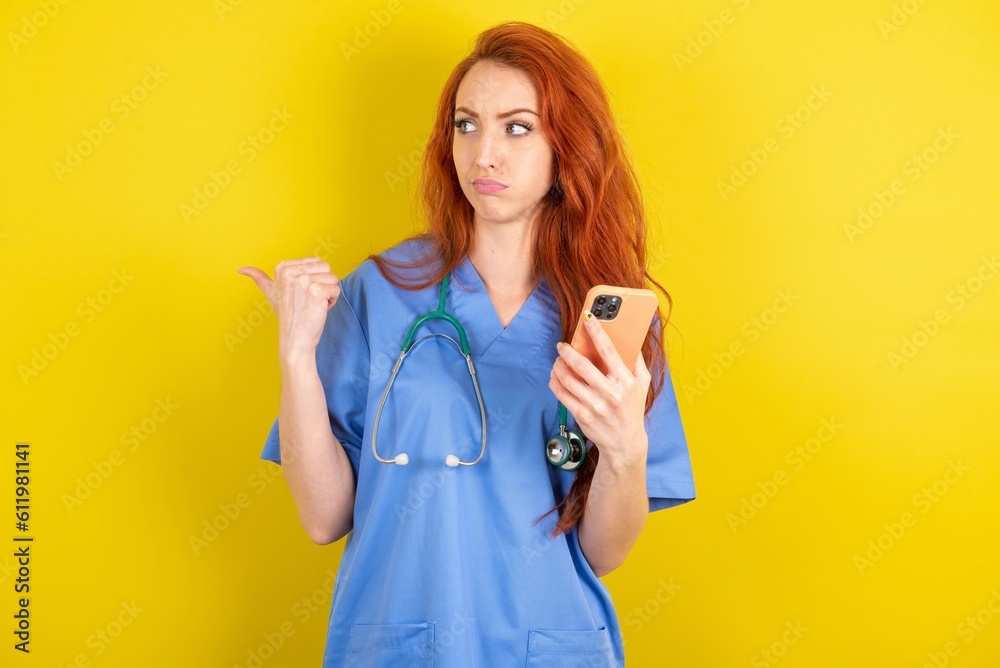 young red-haired doctor woman over yellow studio background points thumb away and shows blank space aside, holds mobile phone for sending text messages. young red-haired doctor woman over yellow studio background points thumb away and shows blank space aside, holds mobile phone for sending text messages.