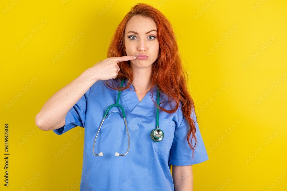Charming young red-haired doctor woman over yellow studio background pointing on pout lips with forefinger, showing effect after lifting procedure, Charming young red-haired doctor woman over yellow studio background pointing on pout lips with forefinger, showing effect after lifting procedure,
